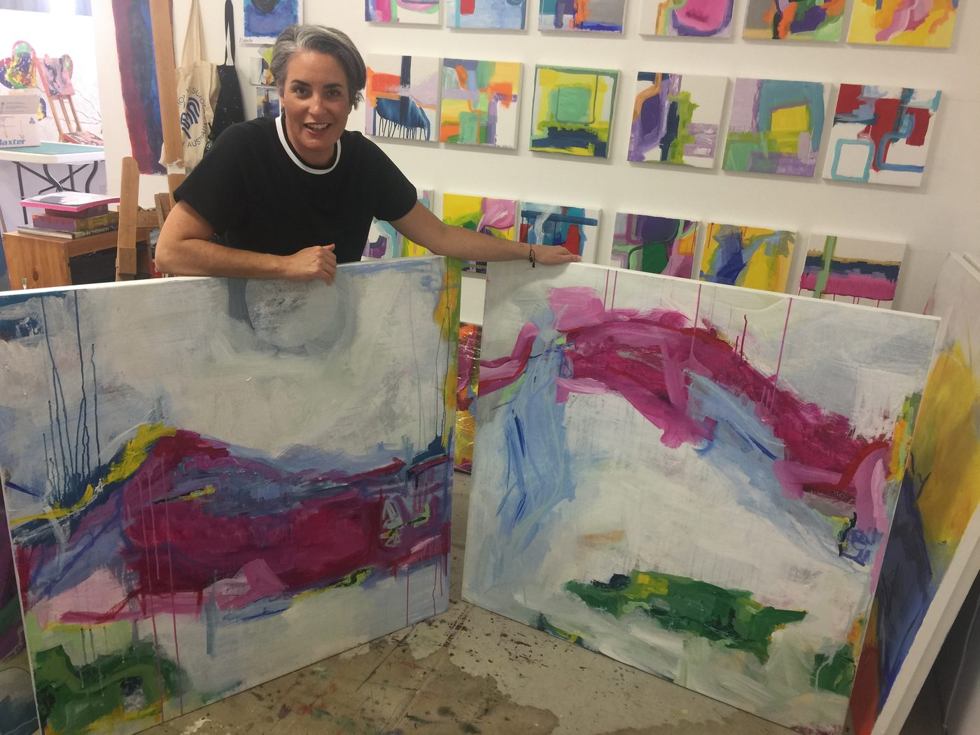 Marisabel González im Atelier hinter zwei großformatigen abstrakten Leinwänden in Pink, Blau und Grün, an der Wand dahinter eine Reihe kleiner farbiger Bilder